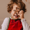 Kind mit lockigem Haar in roter Schürze hebt die Hand – festlicher Moment mit Kinderfreude.